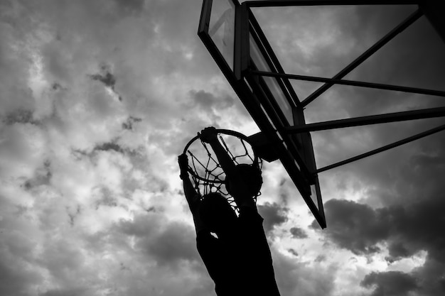 Silhouet van een man die een bal in een basketbalring gooit op straat tegen een lucht met wolken in zwart-witte kleur