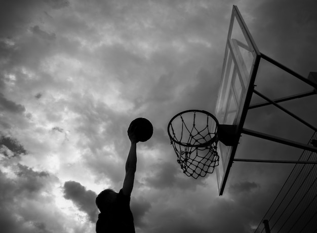 Silhouet van een man die een bal in een basketbalring gooit op straat tegen een lucht met wolken in zwart-witte kleur