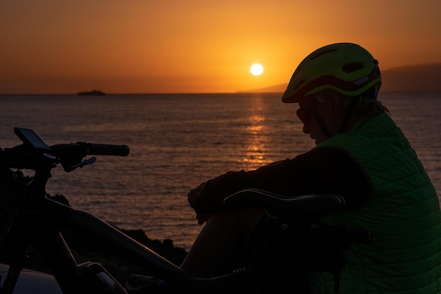 Silhouet van een man die op de klif zit en een beschermende helm draagt na het fietsen langs de zee. oranje-zwarte achtergrond