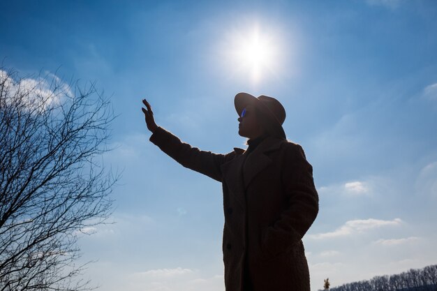 Silhouet van een meisje in een hoed en jas op een achtergrond van de lucht met wolken en de zon.