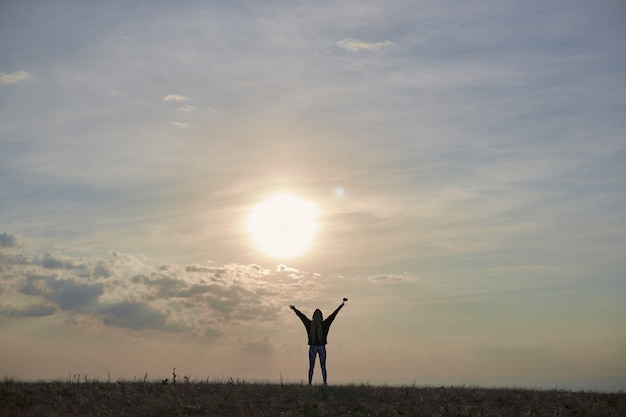 Silhouet van een meisje met lang haar dat op een heuvel staat met haar handen omhoog bij zonsopgang, de zon schijnt...