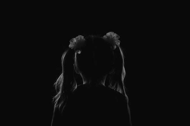 Silhouet van een meisje met twee paardenstaarten die met haar rug in het donkere anonimiteits- en kinderveiligheidsconcept zitten