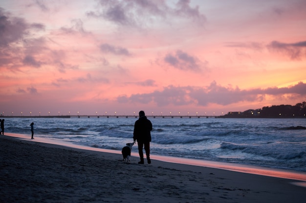 Silhouet van een persoon die op een dag zijn hond op een strand met een rode zonsondergang loopt