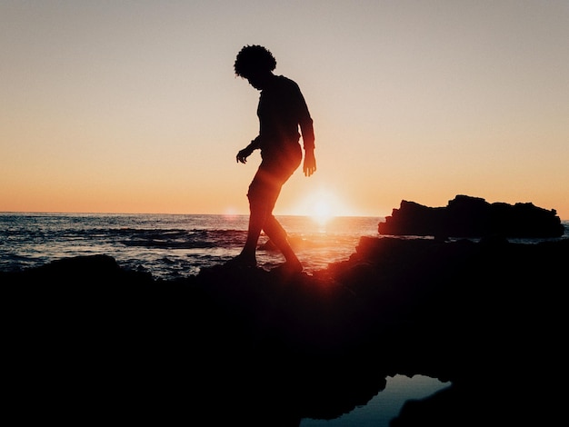 Silhouet van een persoon op een rotsformatie bij de zee tegen een heldere lucht bij zonsondergang