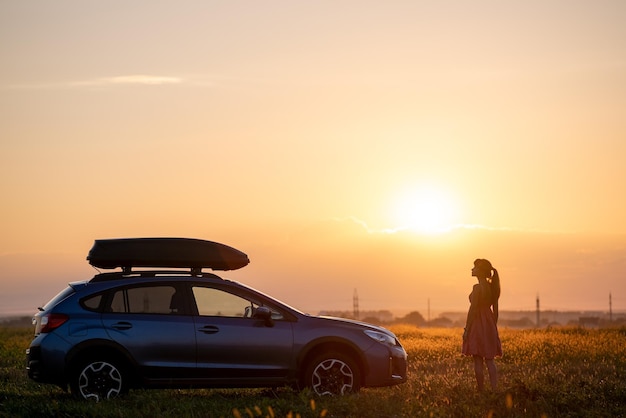 Silhouet van een vrouwelijke chauffeur die in de buurt van haar auto op het grasveld staat en geniet van het uitzicht op de heldere zonsondergang. Jonge vrouw ontspannen tijdens road trip naast SUV-voertuig.