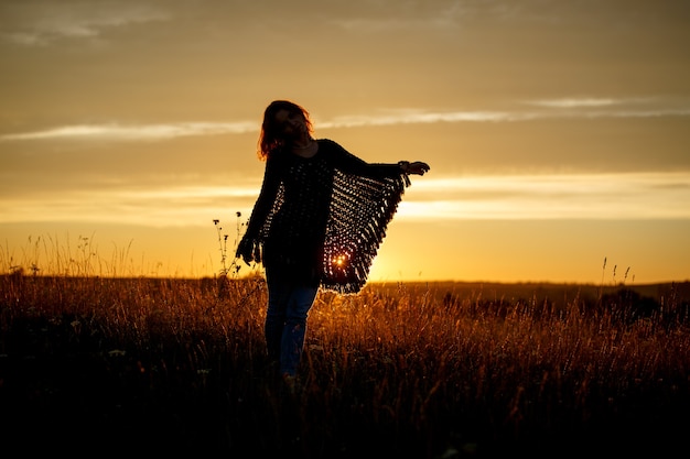 Silhouet van gelukkige jonge vrouw op zonsondergang, buitenmeisje in een geruite poncho in een veld met aartjes