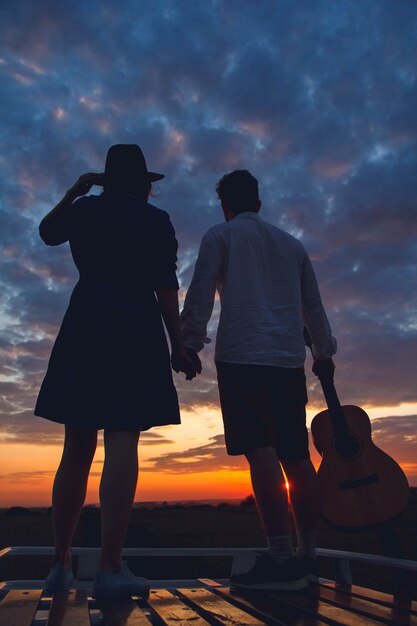 Silhouet van man met gitaar en vrouw in een hoedenstandaard op het dak van een auto op de achtergrond van de zonsondergang