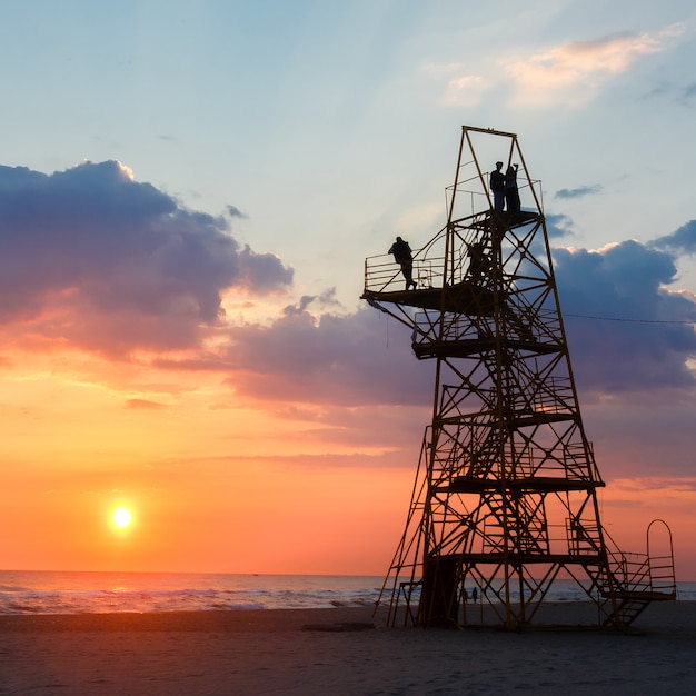 Silhouet van mensen op een reddingstoren op een zandstrand bij zonsondergang.