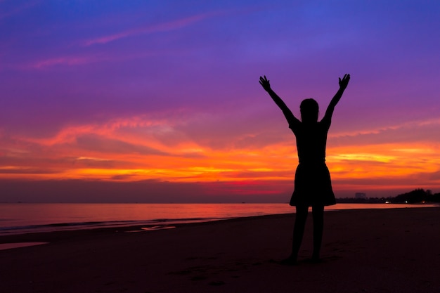 Silhouet van vrouw met handen omhoog terwijl je op zee strand bij zonsondergang