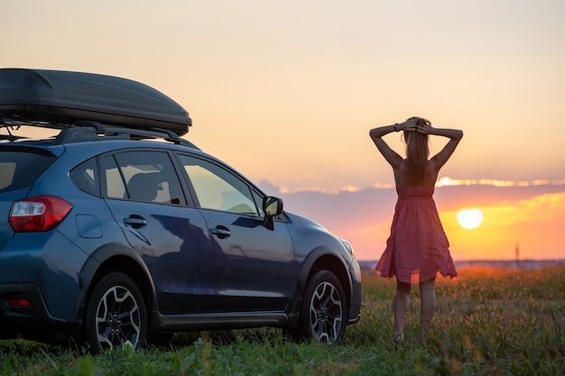 Silhouet van vrouwelijke bestuurder die zich dichtbij haar auto op grasrijk gebied bevindt dat van mening van heldere zonsondergang geniet