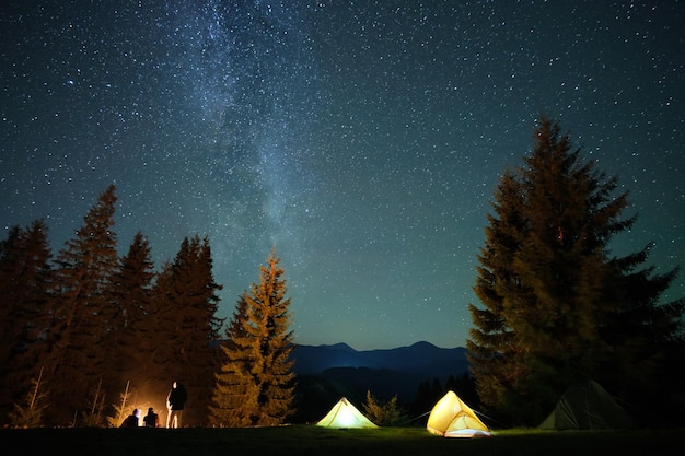 Silhouet van wandelaars die rusten naast het branden van vreugdevuur in de buurt van verlichte toeristententen op de camping in donkere bergen onder de nachtelijke hemel met fonkelende sterren. Actieve levensstijl en buitenleven concept.