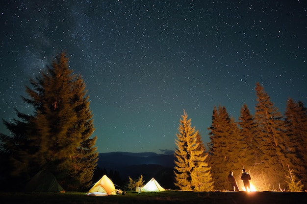 Silhouet van wandelaars die rusten naast het branden van vreugdevuur in de buurt van verlichte toeristententen op de camping in donkere bergen onder de nachtelijke hemel met fonkelende sterren. Actieve levensstijl en buitenleven concept.