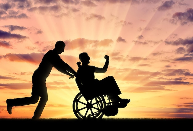 Silhouet vrolijke man met een gehandicapte man in een rolstoel en zijn vriend op een zonsondergang op de achtergrond. Concept van mensen met een handicap in de samenleving