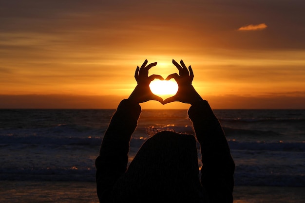 Silhouet vrouw die hartvorm maakt op het strand tegen oranje lucht