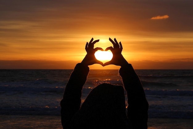 Foto silhouet vrouw die hartvorm maakt op het strand tegen oranje lucht