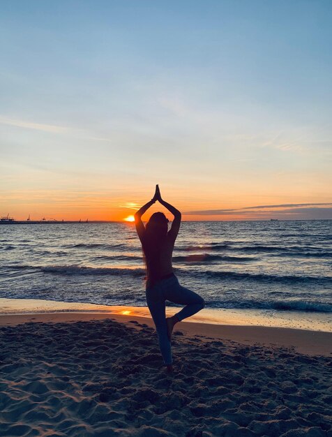 Silhouet vrouw die op één been op het strand staat tegen de hemel tijdens de zonsondergang