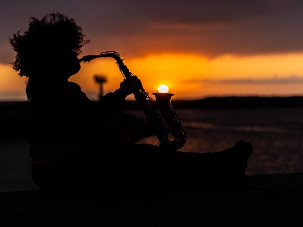 Foto silhouet vrouw die saxofoon speelt zit tegen de hemel tijdens de zonsondergang