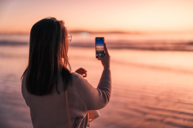 Silhouetportret van een vrouw op het strand bij zonsondergang die een foto maakt met de mobiele telefoon