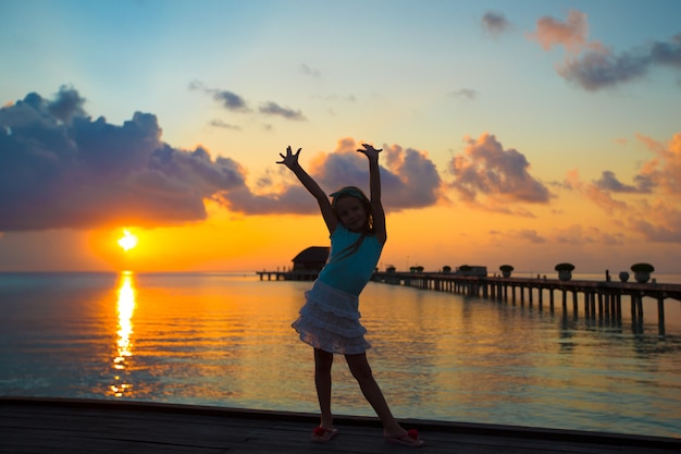 Siluetta della bambina adorabile sul molo di legno al tramonto