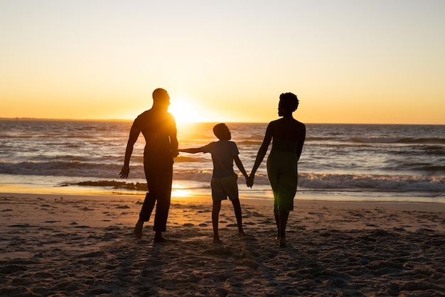 Silhouette african american boy holding mother and father's hands and standing on beach at sunset. Copy space, unaltered, sea, sky, family, together, childhood, nature, vacation, enjoyment, scenery.
