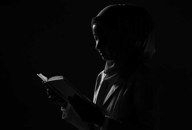 Silhouette Asian muslim girl in hijab dress is praying with a book on black background