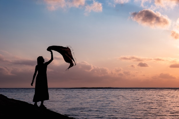 Profili la donna asiatica che si rilassa con alzato il panno sul mare tropicale con il cielo variopinto