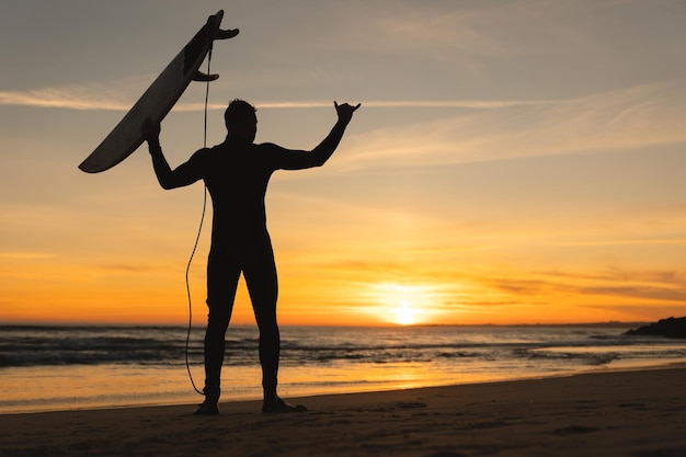 Silhouette of an athletic man standing on the seashore holding a surfboard and showing shaka at suns