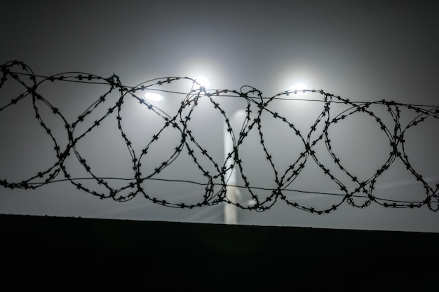 Silhouette of barbed wire on a fence against a background of\
blurry lamp posts at foggy night