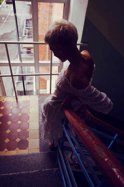 Silhouette of a blonde woman in a white semitransparent dress in a semidark entrance on the stairs