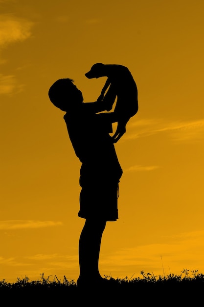 写真 夕暮れの空に向かって畑に立っている子犬を背負っているシルエットの男の子