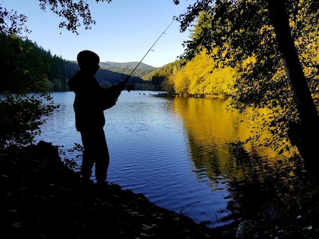 Silhouette boy fishing in lake against clear sky