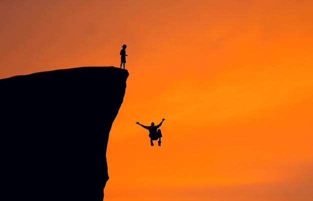Silhouette boy looking at man falling from cliff against clear orange sky