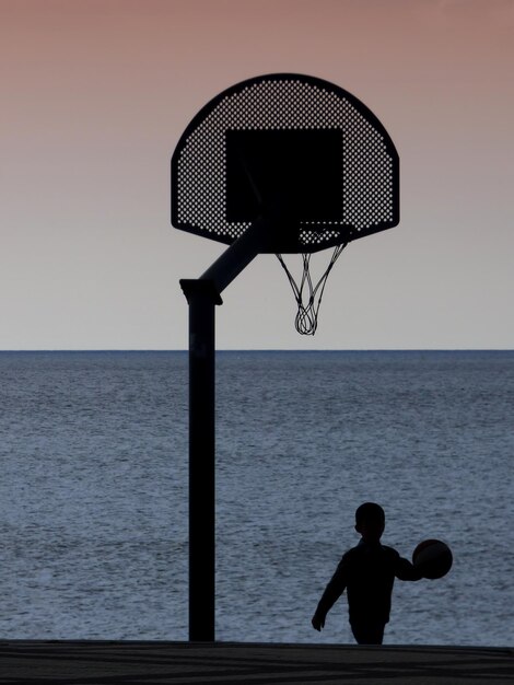 Photo silhouette boy playing basketball by sea during sunset