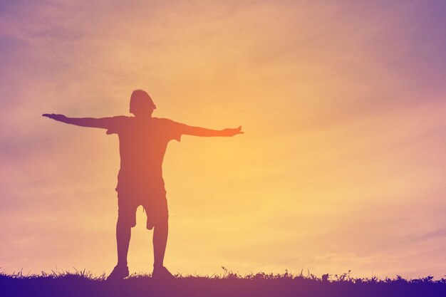 Silhouette boy with arms outstretched standing on field against cloudy sky during sunset