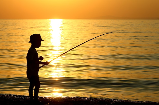 Silhouette of a boy with a fishing rod on sea in Greece in summer