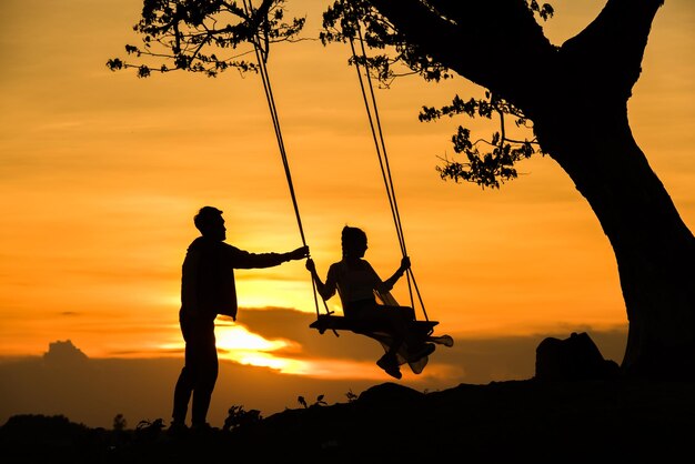 Silhouette boyfriend swinging girlfriend on rope swing against orange sky during sunset