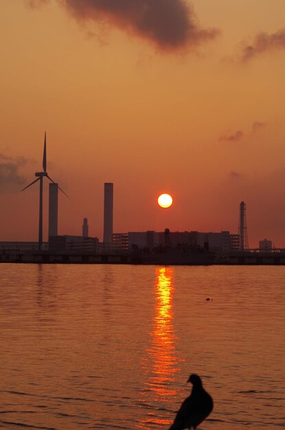 Silhouette buildings by sea against orange sky