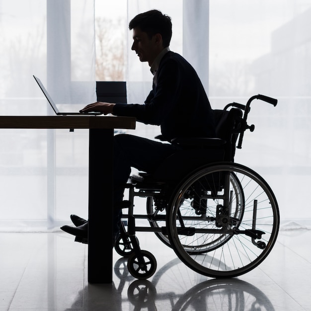 Photo silhouette of a businessman sitting on wheelchair using laptop on table