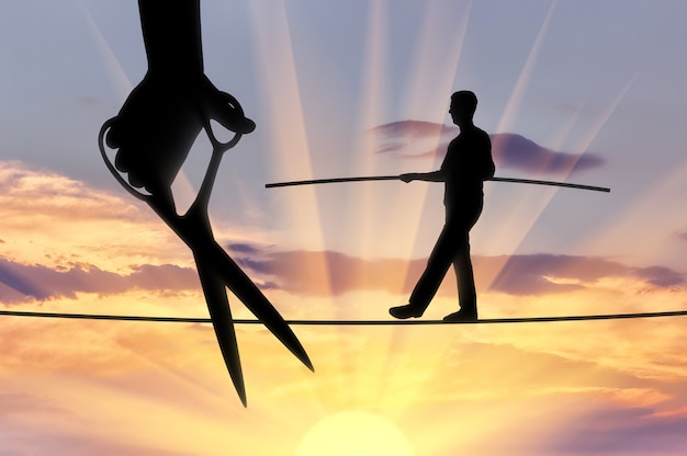 Silhouette of a businessman walking on a tightrope balances and a hand with scissors intends to cut the rope. The concept of meanness in business