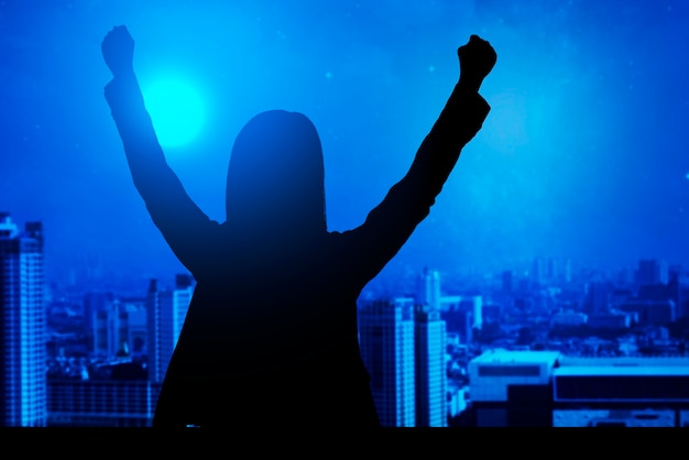 Silhouette of a businesswoman standing and raised hands with cityscapes background