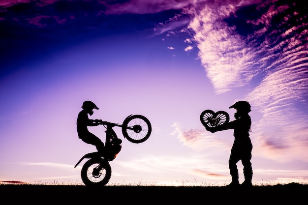 Силуэт пара стоял и сердце форму в руке с мотоциклом на закате.