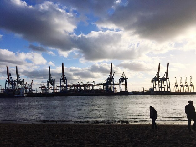 Silhouette cranes at commercial dock against cloudy sky