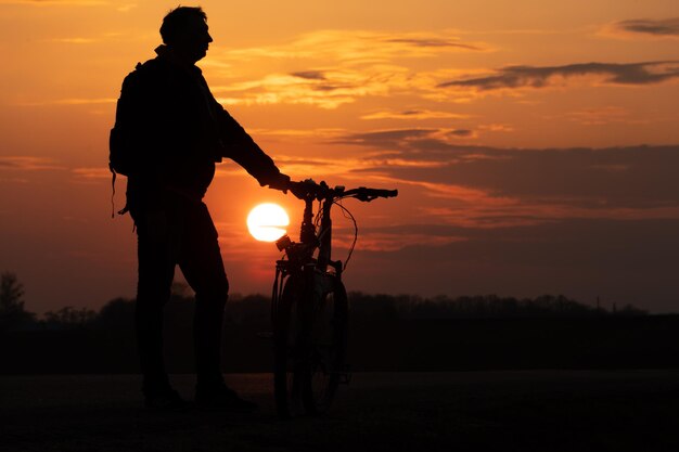 太陽と美しい空を背景にしたサイクリストのシルエット 夕日を背景に自転車の隣に立っている男の輪郭