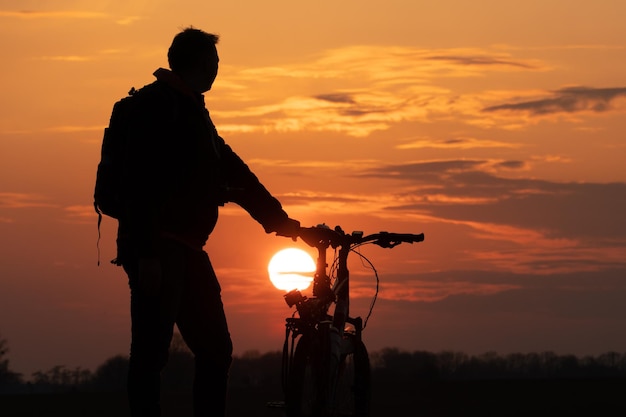 太陽と美しい空を背景にしたサイクリストのシルエット 夕日を背景に自転車の隣に立っている男の輪郭