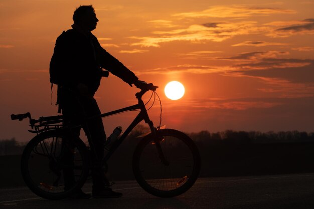 太陽と美しい空を背景にしたサイクリストのシルエット 夕日を背景に自転車の隣に立っている男の輪郭