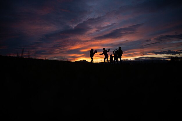 日没時の丘の外でボールで遊ぶ家族のシルエット