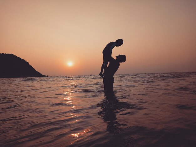 Photo silhouette father carrying son in sea against sky during sunset