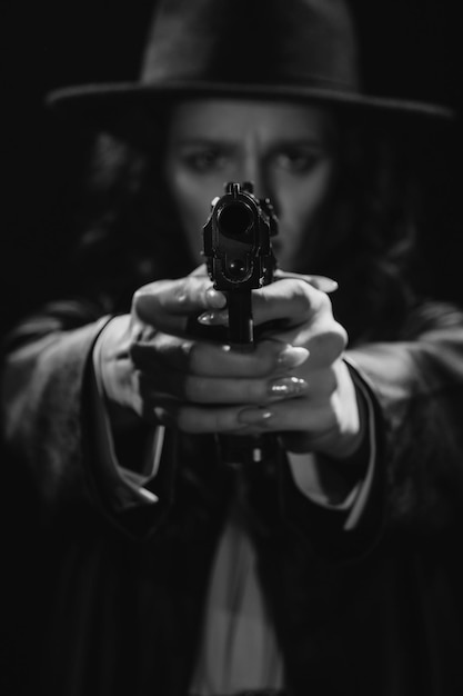 Силуэт женщины-детектива в пальто и шляпе с пистолетом в руках, указывающим на камеру Книжный драматический нуар-портрет в стиле детективов 1950-х черно-белый снимок