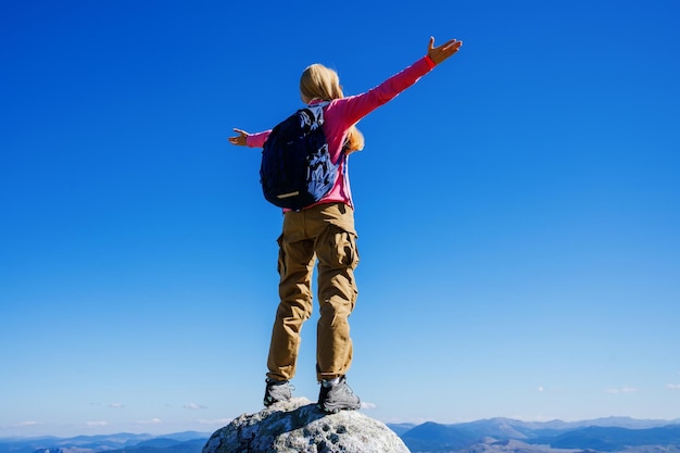 崖の上に立って山頂を楽しむバックパックを持った女性観光客のシルエット 青い空と彼女の前の山々