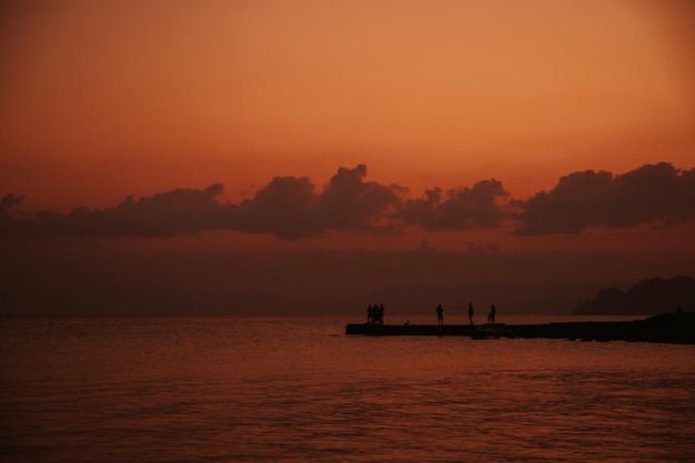 Silhouette of fishermen fishing on pier in sea at sunset against background of mountains